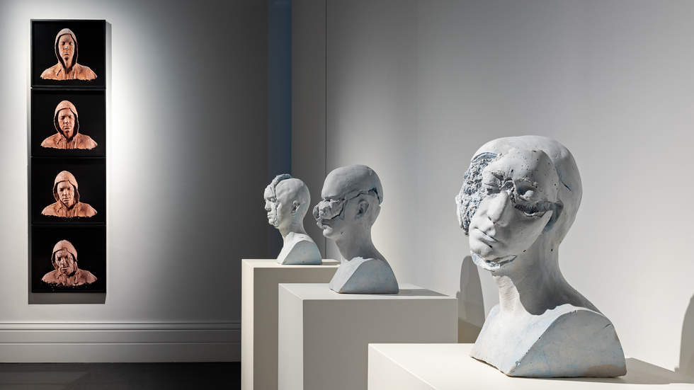 A gallery interior with sculpture busts on plinths and photographs of terracotta heads hung on wall.