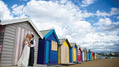 Married couple kissing in front of multi-coloured beach bathing boxes.