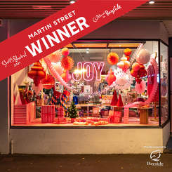 Glass shopfront with pink illuminated Joy sign and a range of red decorations including presents and giant baubles.