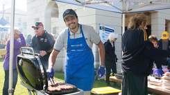 A man wearing a blue apron standing over a bbq. 