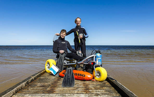 Disabled diver sitting on equipment and person standing at edge of water at beach