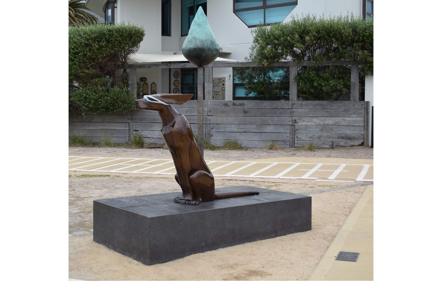 View of an outdoor bronze sculpture of an elongated dog leaning forward and wears sunglasses. 
