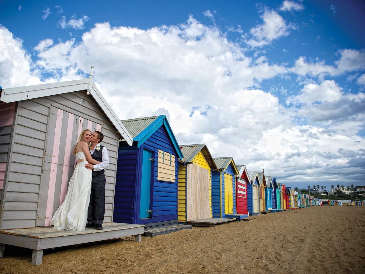 Married couple kissing in front of multi-coloured beach bathing boxes.