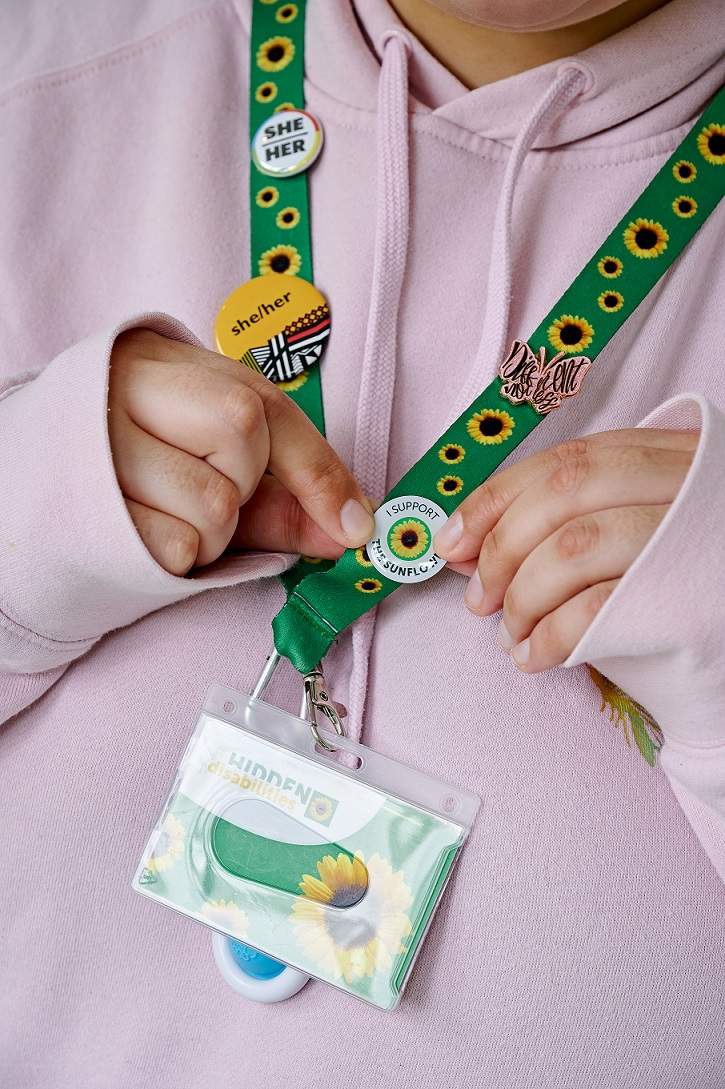 Sunflower lanyard with four buttons. Two buttons read she/her, one button reads divergent and the last button is being held and reads I support the sunflower