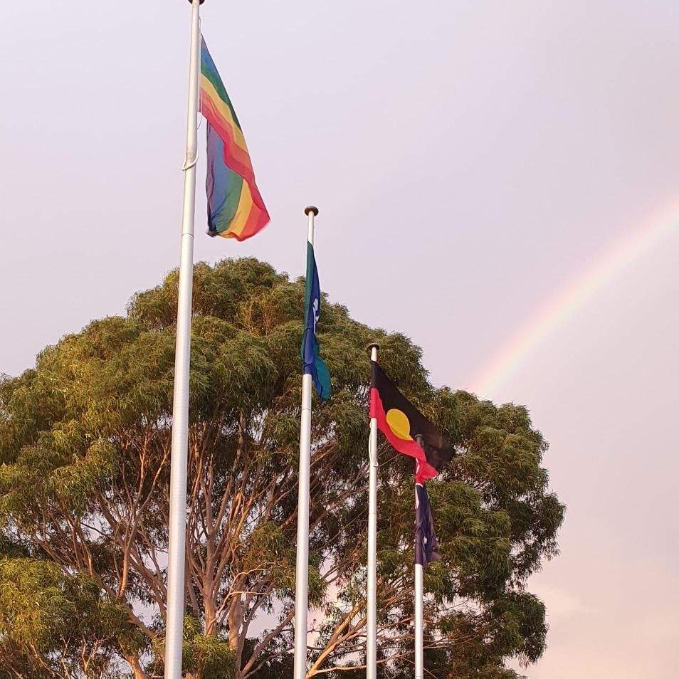 Four flags outside Bayside City Council's offices with a rainbow in the sky behind them.