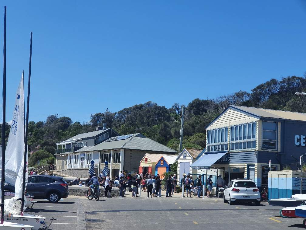 Image of people outside Cerberus Cafe at Half Moon Bay, Black Rock. Boatsheds and Half Moon Bay Life Saving Club in background.