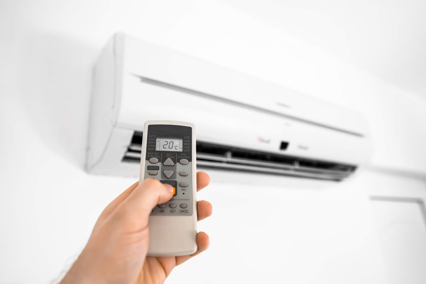 A hand holding an air conditioner remote, with a reverse cycle air conditioner on a white wall in the background