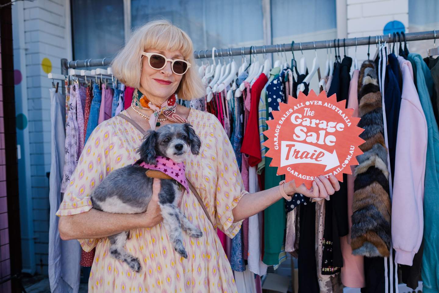 A blonde woman holding a small grey dog and a red Garage Sale Trail sign, standing in front of a full clothing rack