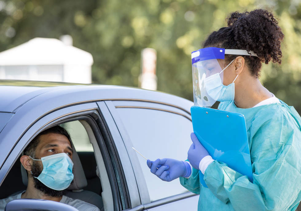 Health worker in full PPE with clipboard and swab talks to a man in a car