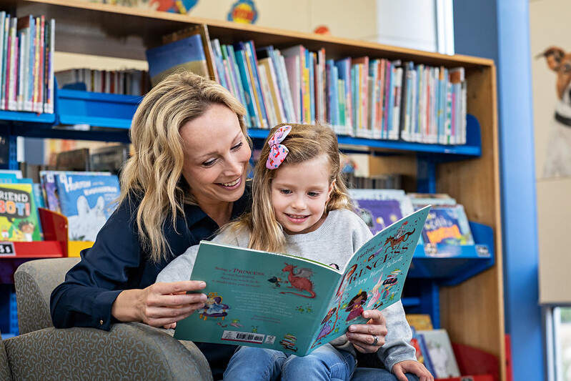 A woman reads to a little girl on her lap with a shelf of books behind them.