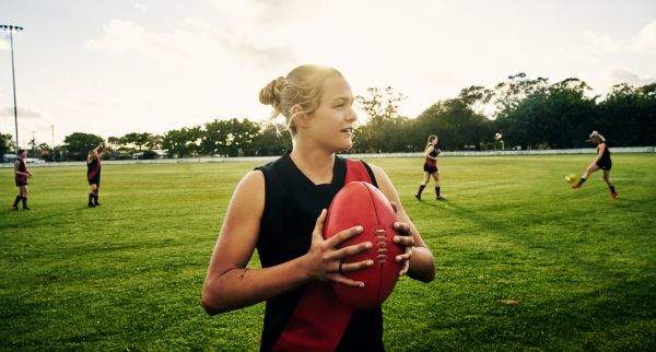 Female footballer at an oval holding a football with players in the background