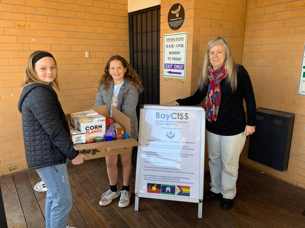 Two young people holding box of food with another person standing next to a sign